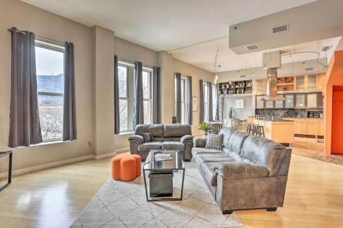Spacious living area with gray sofas, orange ottoman, large windows, and an open kitchen in a modern apartment.