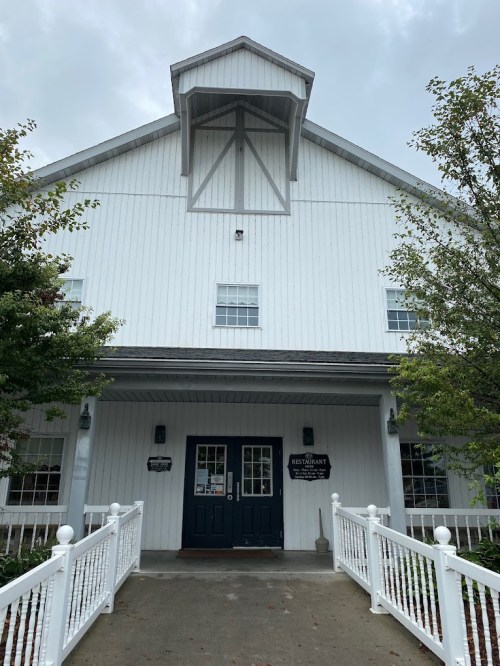 A white barn-style building with a dark door and windows, surrounded by greenery and a white railing.