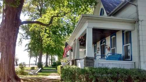 A charming house with a porch, American flag, and lush greenery, overlooking a serene waterfront.