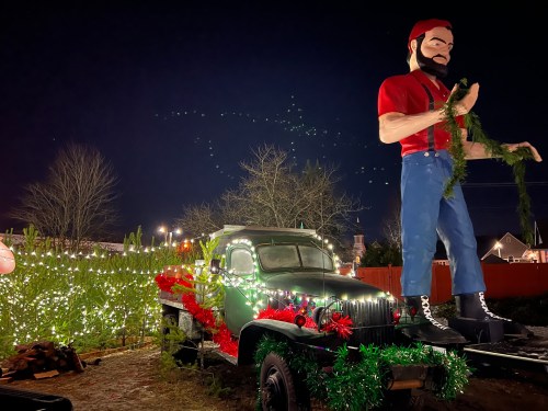 A large statue of a man in a red shirt stands on a decorated vintage truck, surrounded by festive lights and greenery at night.