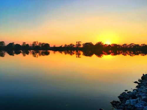 A serene sunset over a calm lake, reflecting vibrant orange and yellow hues with silhouettes of trees in the background.