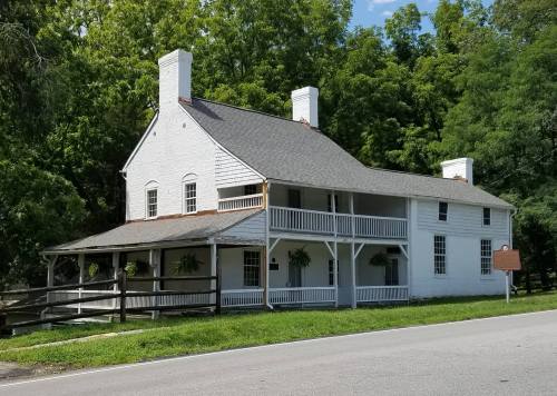 Historic white house with a porch, surrounded by greenery, located near a road.
