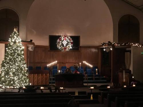 A warmly lit church interior decorated for the holidays, featuring a Christmas tree, wreath, and candles.