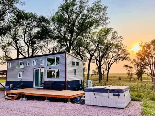 A modern tiny house with a wooden deck and hot tub, surrounded by trees at sunset.