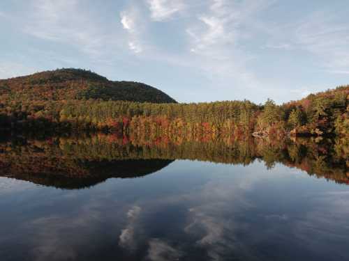 A serene lake reflecting colorful autumn trees and a mountain under a blue sky with wispy clouds.