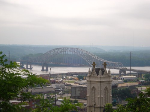 A view of a bridge spanning a river, with a cloudy sky and a church steeple in the foreground.