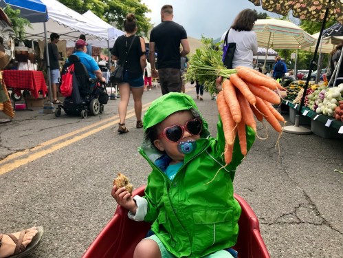 A child in a green jacket and sunglasses holds up fresh carrots while sitting in a wagon at a bustling farmers market.