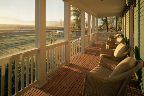 A serene porch with wicker chairs overlooks a misty landscape at dawn, surrounded by greenery and a wooden fence.