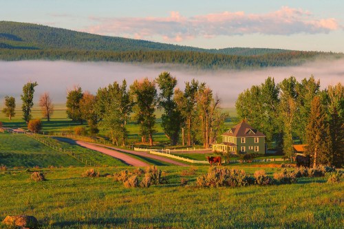 A serene countryside scene with a house, trees, and a misty landscape under a colorful sky at sunrise.