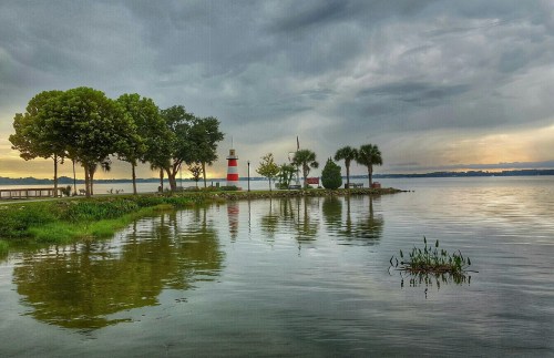 A serene lakeside scene featuring a lighthouse, trees, and reflections on the water under a cloudy sky.