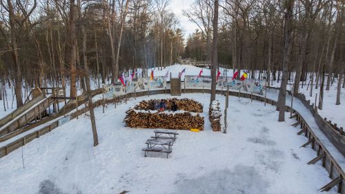 A snowy forest scene with a circular stack of firewood and flags, surrounded by trees and a wooden pathway.