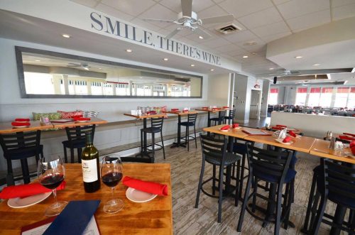 A bright restaurant interior with wooden tables, black chairs, and a wine bottle on the table, featuring a cheerful sign.