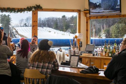 A cozy bar scene with patrons enjoying drinks, overlooking a snowy slope through large windows.