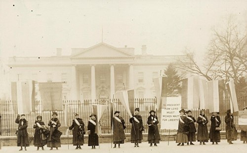 Women in long coats and sashes protest outside the White House, holding banners for women's rights and liberty.