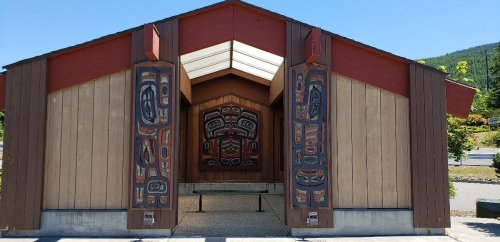 A traditional building with totem pole carvings on the entrance, surrounded by greenery and mountains in the background.