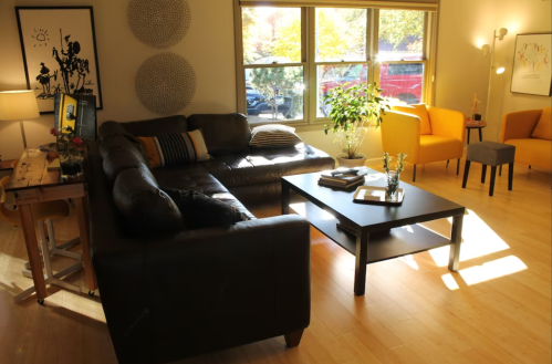 Cozy living room with a black sectional sofa, yellow chairs, a coffee table, and large windows letting in natural light.