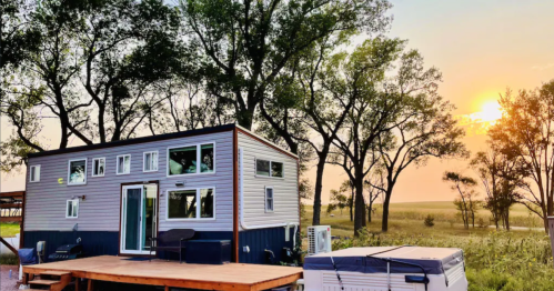 A modern tiny house with large windows, surrounded by trees, at sunset in a serene outdoor setting.
