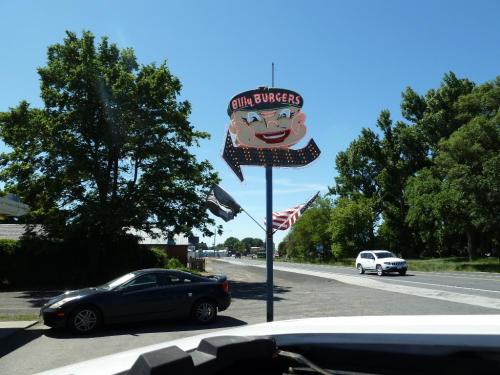 Sign for "Billy Burgers" featuring a smiling cartoon chef, with flags and trees in the background on a sunny day.