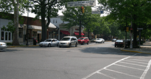 A busy street scene with cars, trees, and storefronts under a clear sky. Traffic lights and signs are visible.