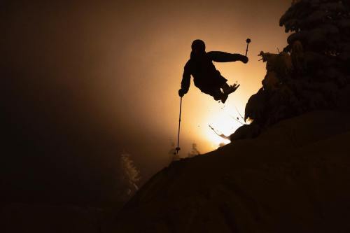 A silhouetted skier jumps against a glowing background of snow and trees, capturing a dynamic winter scene.
