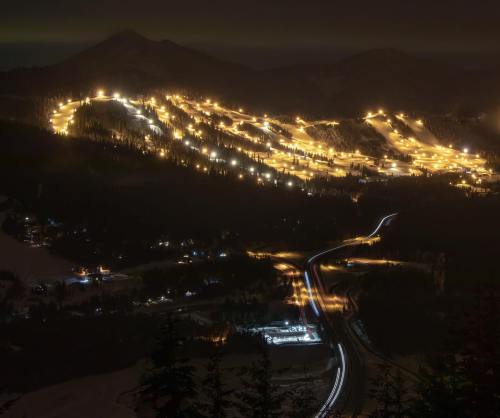 A nighttime view of a ski resort, illuminated trails winding down a mountain, with glowing lights and dark surroundings.