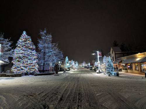 Snow-covered street at night, lined with colorful Christmas lights and decorated trees, creating a festive winter scene.