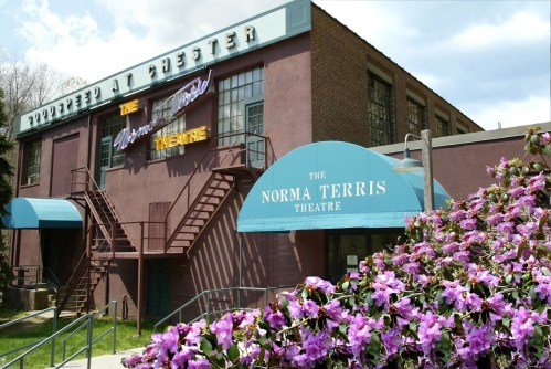 Exterior of the Norma Terris Theatre with blue awnings and blooming flowers in the foreground under a partly cloudy sky.