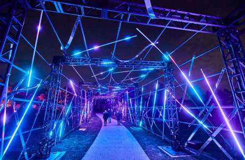 A vibrant pathway illuminated by blue and purple lights, with people walking through a geometric light installation at night.