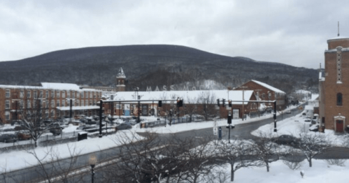 Snow-covered town with buildings and a mountain in the background, under a cloudy sky. Traffic lights visible.
