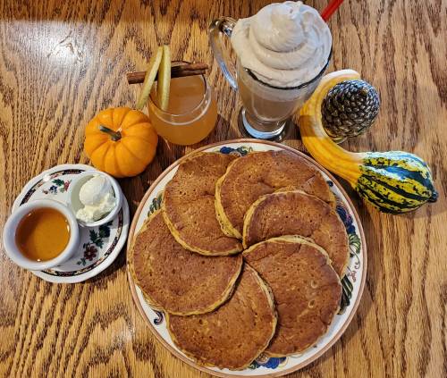 A plate of pancakes with syrup and whipped cream, alongside a pumpkin, drink, and decorative gourds on a wooden table.
