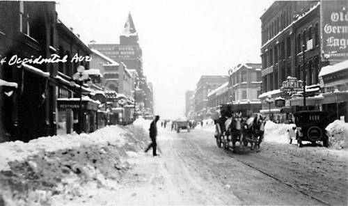 Historic snowy street scene with horse-drawn carriages and pedestrians, lined with buildings and storefronts.