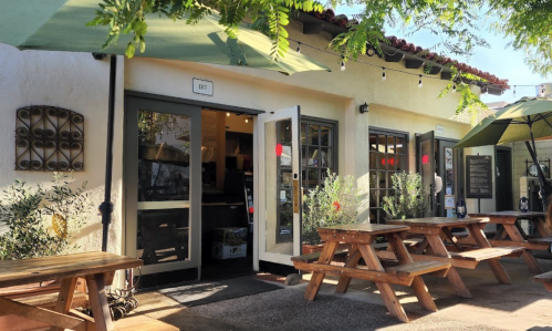Outdoor seating area with wooden tables and umbrellas in front of a café entrance, surrounded by greenery.