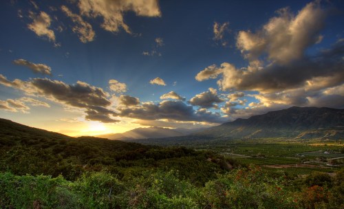 A scenic landscape at sunset, featuring mountains, lush greenery, and dramatic clouds in a colorful sky.