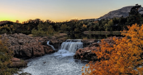 A serene waterfall cascades over rocks, surrounded by autumn foliage and a tranquil lake at sunset.