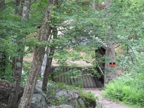 A wooden covered bridge nestled among trees, with a warning sign visible near the entrance.