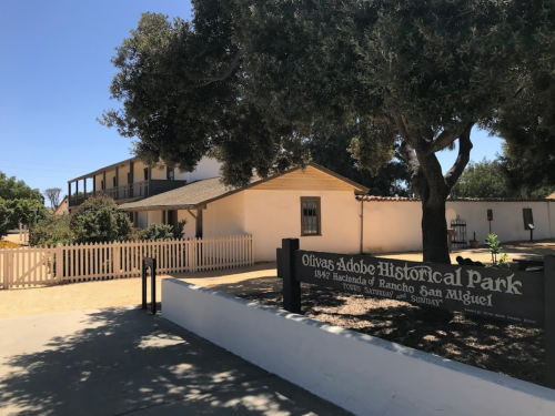 Olivas Adobe Historical Park sign in front of a historic building surrounded by trees and a white fence.