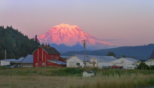 A scenic view of a snow-capped mountain at sunset, with a red barn and white houses in the foreground.