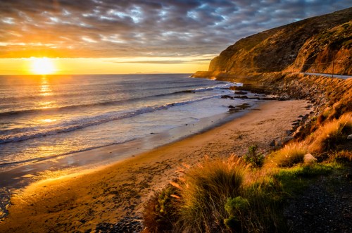 Sunset over a serene beach with gentle waves, rocky cliffs, and lush grass in the foreground.