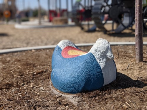 A colorful rock sculpture painted with the Colorado state flag, set in a playground area with mulch and equipment in the background.