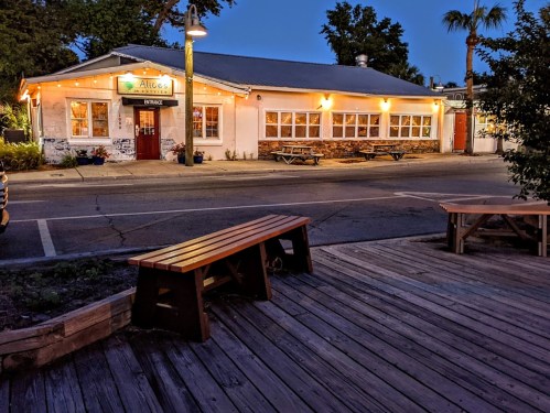 A cozy restaurant exterior at dusk, with warm lights and outdoor seating, surrounded by trees and a quiet street.