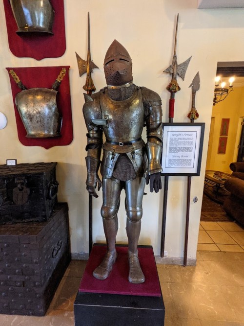 A full suit of medieval armor displayed on a stand, with weapons and armor pieces on the wall behind it.