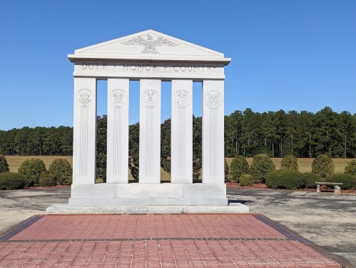 A stone monument with four columns, inscribed "DUTY, HONOR, COUNTRY," set against a clear blue sky and green trees.