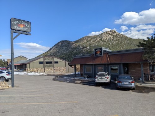A restaurant with a sign reading "Smokin' Dave's" next to a mountain, with parked cars and a clear blue sky.
