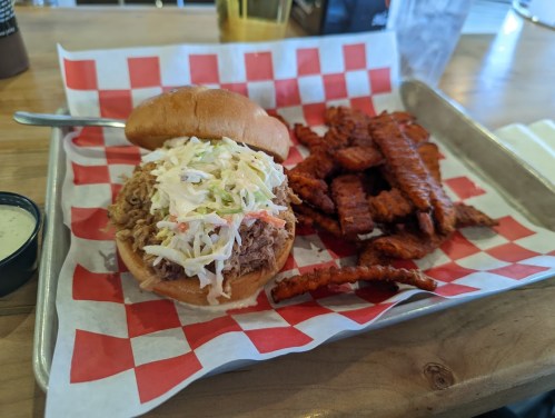 A pulled pork sandwich with coleslaw on a bun, served with sweet potato fries on a checkered paper-lined tray.