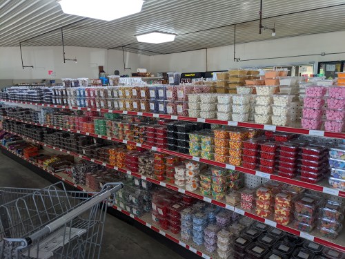 A colorful display of various candies and sweets arranged neatly on shelves in a store. A shopping cart is visible in the foreground.