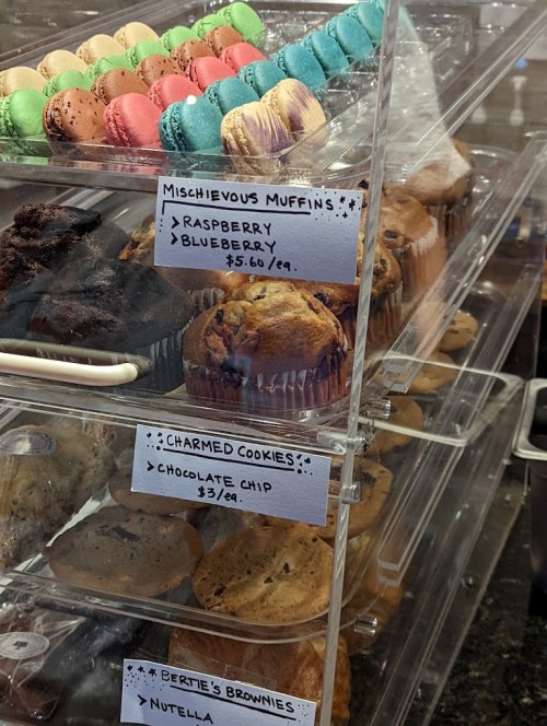Display case with colorful macarons, muffins, cookies, and brownies, labeled with prices and flavors.