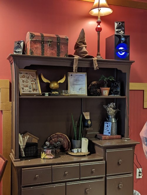 A wooden shelf displays a mix of decorative items, including a treasure chest, plants, and a lamp, against a red wall.