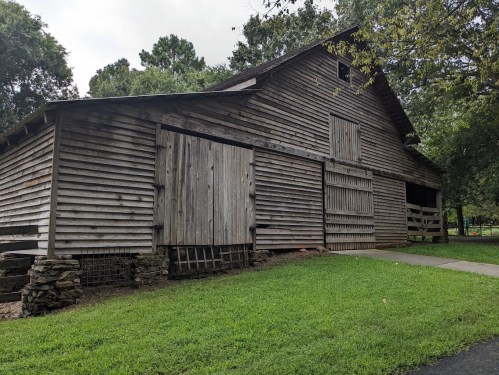 A weathered wooden barn with a sloped roof, surrounded by green grass and trees, on a cloudy day.