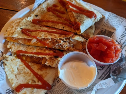 A plate of quesadillas drizzled with sauce, served with salsa and sour cream on a bed of newspaper.