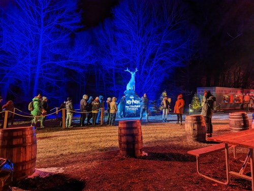 A nighttime scene with blue lighting, featuring a line of people near a statue and wooden barrels in a festive outdoor setting.
